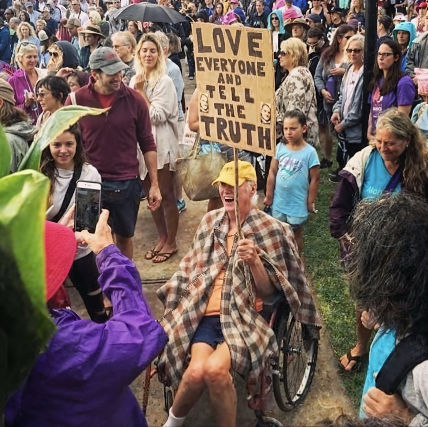 A person in a crowd holding a handmade sign reading Love everyone and tell the truth.